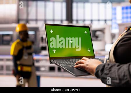 African american factory worker holds laptop with green screen display, overseeing automated production line. The industrial plant integrates robotics, conveyor belts and smart manufacturing systems. Stock Photo