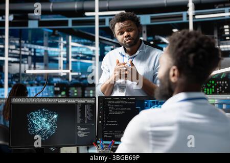 Server farm manager supervising engineer looking at data analysis graph to reduce failures. Data center team leader overseeing worker looking at AI visualization dashboard to pinpoint gear glitches Stock Photo