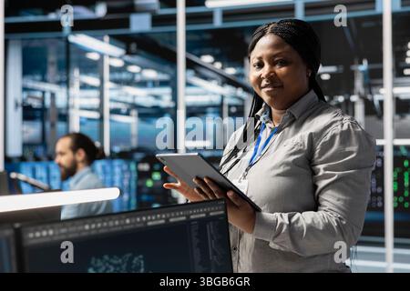 Portrait of smiling data center programmer using tablet to update servers tech, configuring gear using software tools. Happy server farm worker reviewing hardware analytics using device Stock Photo