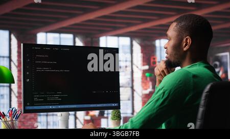 Black technician in home office looks at UI on computer monitor, typing and executing programming code. IT worker using coding application to write and test code in modern apartment. Camera A. Stock Photo