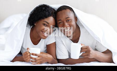 Happy black couple laying under blanket, drinking tea Stock Photo