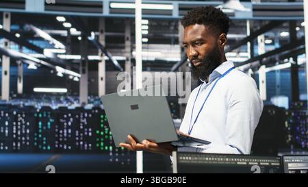 Data center programmer uses node tree software to identify equipment performance issues. African american server farm worker reviews data analysis graph to minimize hardware failures, camera B Stock Photo