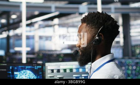 Data center employee talking in headset mic, coordinating technicians setting up hardware for artificial intelligence deployment. Server room worker engages in remote call with engineers, camera B Stock Photo