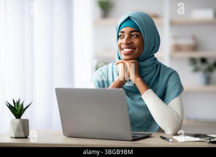 Smiling Young Black Islamic Woman In Hijab Sitting At Desk With Computer Stock Photo