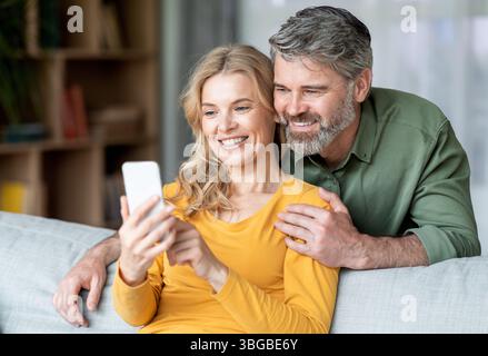Portrait Of Smiling Middle Aged Couple Resting With Smartphone On Couch Stock Photo