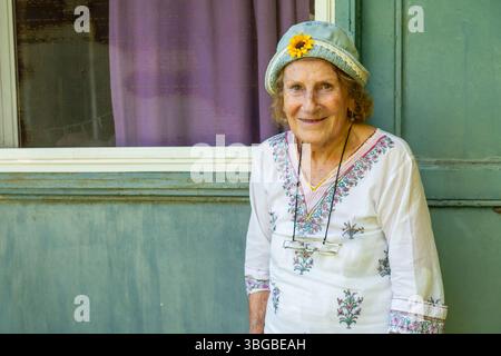 Elderly woman smiling while leaning against a light green wooden door, wearing a light blue hat with a sunflower and a white floral-patterned shirt by Stock Photo