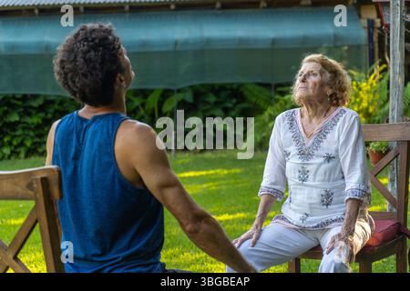 Senior woman and her physiotherapist are sitting on chairs in a garden, practicing breathing exercises together, focusing on improving respiratory hea Stock Photo