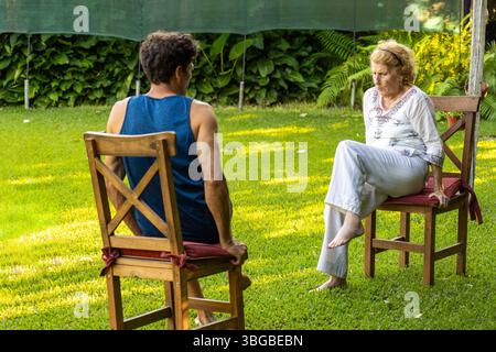 Senior woman and her physiotherapist are sitting on chairs in a garden, practicing rehabilitation exercises to improve mobility and well-being Stock Photo