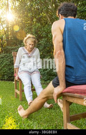 Senior woman exercising her legs with the help of a personal trainer, sitting on chairs in a backyard with green grass and trees, during a sunny day Stock Photo
