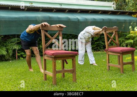 Elderly man and woman practicing stretching exercises with chairs on a grassy lawn in a serene garden setting, promoting health and flexibility in old Stock Photo