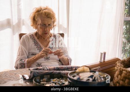Concentrated senior seamstress meticulously threading a needle, surrounded by sewing materials on a table at home, illuminated by natural light Stock Photo
