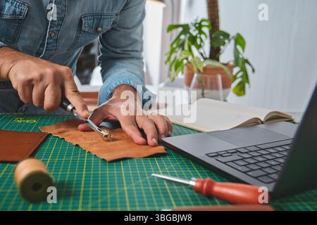Craftsman working leather using stitching wheel in his workshop with laptop, notebook and tools Stock Photo