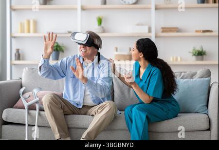Disabled senior man using VR headset with young nurse, exploring augmented reality at home Stock Photo