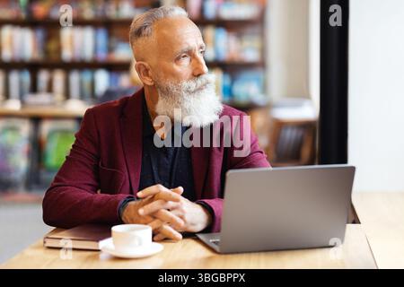 Pensive grey-haired businessman using laptop at cafe, looking through window Stock Photo