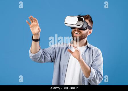 Happy bearded young man using VR headset to explore augmented reality on blue studio background Stock Photo