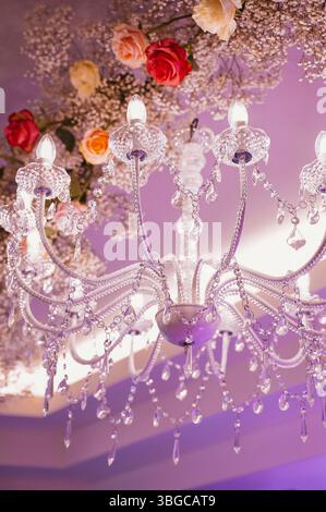 Crystal chandelier draped with delicate flowers and sparkling crystals. Soft pink and warm orange hues of roses enhance the luxurious elegance of the Stock Photo