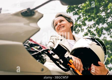 Confident gray-haired woman holding helmet beside motorcycle, symbol of freedom and graceful aging Stock Photo