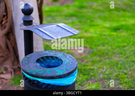 Detailed view of a weathered black metal trash bin with a blue liner and canopy lid, placed on green grass in a park setting. Stock Photo