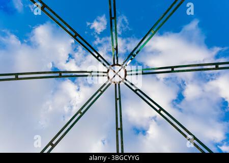 Symmetrical green metal structure viewed from below with a circular center, contrasted against a vibrant blue sky with scattered clouds. Stock Photo