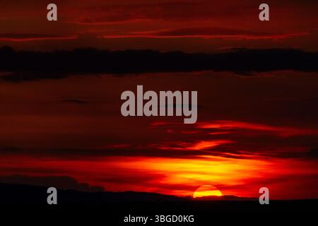 Close-up of a glowing orange sunset with sun half-hidden behind distant hills and dark dramatic clouds Stock Photo
