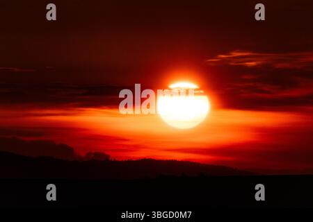 Vibrant round sun setting behind silhouetted ridges, bathing the sky in fiery tones of red and orange for a stunning sunset moment Stock Photo