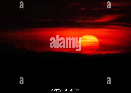 Low on the horizon, the sun sinks into a bed of glowing clouds, painting the sky in layers of red and orange above the distant hills Stock Photo