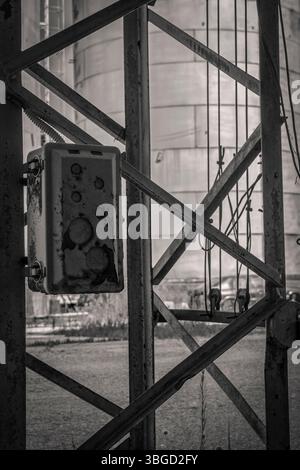Close-up of a weathered electrical box attached to a metal framework, with an industrial silo in the background, conveying a sense of decay and abando Stock Photo