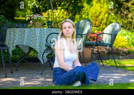 A blonde woman sits cross-legged on the edge of a garden patio, looking thoughtfully into the distance. Stock Photo