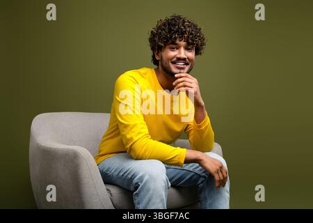Cheerful young man with curly black hair sitting on a gray chair wearing a yellow shirt with khaki background Stock Photo
