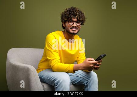 Young man with curly hair smiling while using a smartphone, sitting casually on a modern chair against a khaki background Stock Photo