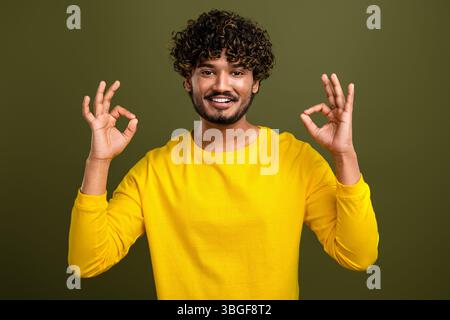 Young curly haired man in yellow shirt making OK gesture with both hands against khaki background Stock Photo