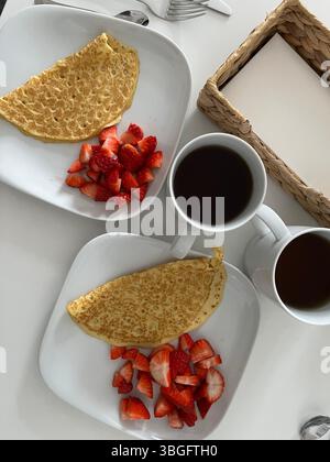 Minimalist breakfast setting with two white plates of crepes and fresh strawberries, served alongside black coffee in white mugs on a white table. Stock Photo