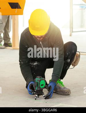 Construction worker wearing protective workwear and yellow hardhat using rotating laser level on tripod, taking measurements at construction site with bright sunlight Stock Photo
