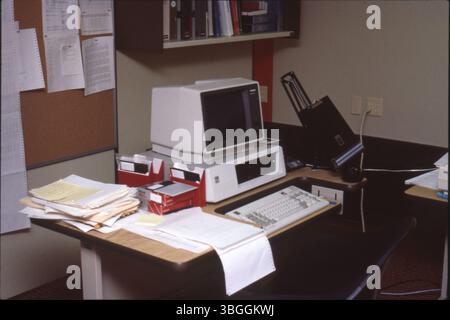 A personal computer sits on a desk in an office, circa 1985. The setup includes a monitor, hard drive, and keyboard. Nearby, there are boxes of 5 1/4-inch floppy disks and a stack of papers. Stock Photo