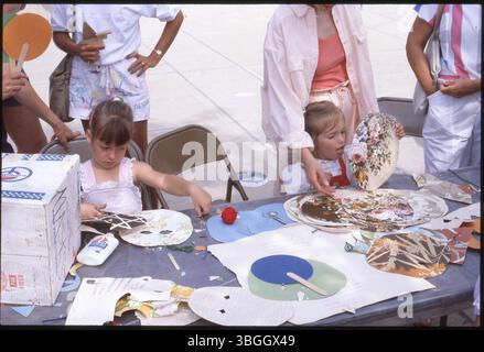 Two children sit at a table working on art projects during an unknown event. They appear unhappy, with adults standing behind them. Stock Photo