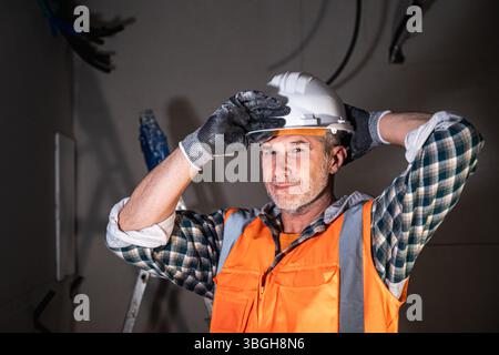 Experienced male construction worker adjusting his safety helmet on-site, wearing a high-visibility vest, gloves, and plaid shirt inside an unfinished Stock Photo