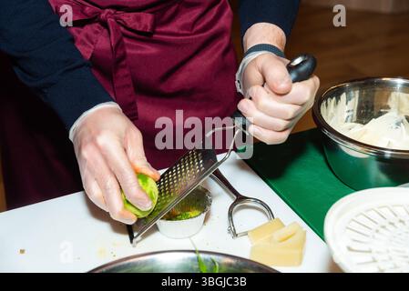 Close-up of gloved hands zesting a lime over a metal grater, with cheese, greens, and cooking tools on the counter. Stock Photo