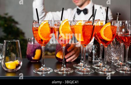 Row of vibrant orange cocktails with orange slices and straws on a bar counter, with bartender preparing drinks in background. Stock Photo