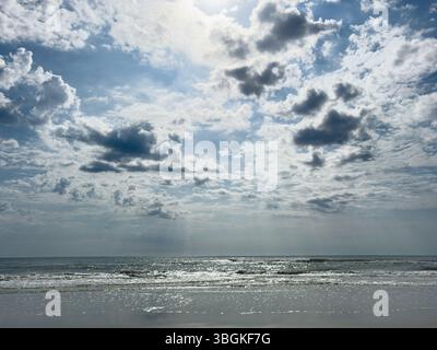A powerful midday scene of sunlight piercing through dramatic clouds above a tranquil ocean shore. This high-resolution image captures the contrast be Stock Photo