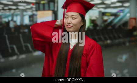Woman in graduation cap and gown standing in airport terminal, wearing red academic robe, symbolizing achievement and transition, surrounded by blurre Stock Photo