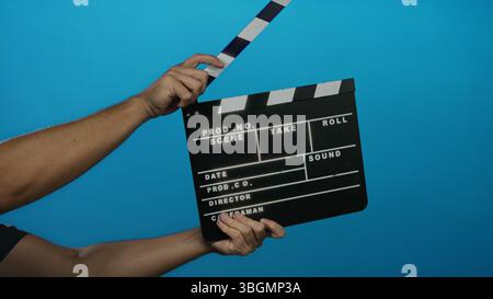 Male hand holding clapperboard against blue background ready for action in film production scene. Stock Photo