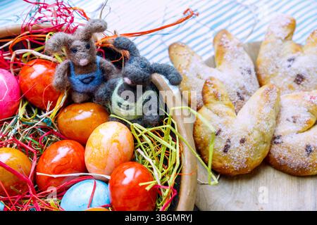 Colored Easter eggs in an Easter nest with colorful natural raffia with felted Easter bunnies and plate with curd Easter bunnies on a striped tablecloth Stock Photo
