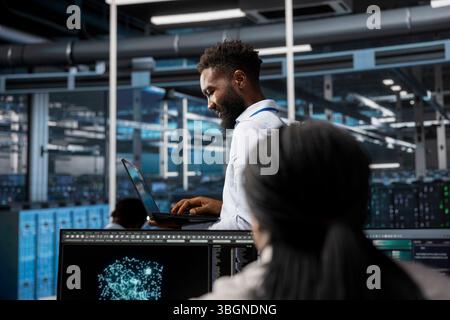 Data center system administrator uses AI visualization tech to oversee automated repetitive tasks. African american man in server farm modifying infrastructure rigs based on data analytics graphs Stock Photo