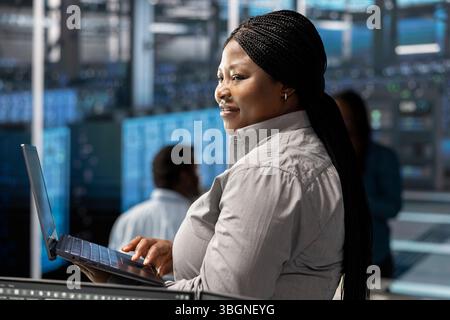 Smiling technician in server room using laptop to implement and manage data backup solutions. Happy data center engineer identifying and resolving technical issues, ensuring efficient recovery Stock Photo