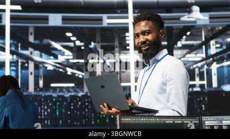 Happy data center admin uses node tree programming on laptop to identify equipment performance issues. Cheerful server farm worker reviews data analysis graph to minimize hardware failures, camera B Stock Photo