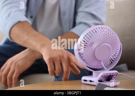 Young man turning mini fan on table at home, closeup Stock Photo