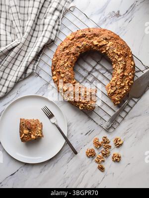 Top down view of bundt cake with walnuts cooling on a rack with a slice on a plate, on a marble surface Stock Photo