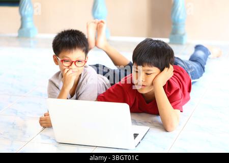 Two children lie on the floor, focused on a laptop in a bright room, showcasing a moment of learning and interaction through technology and friendship Stock Photo