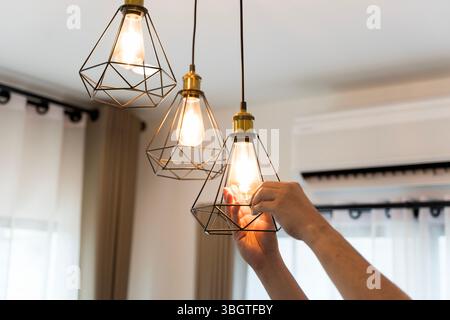 Close-up of a hand replacing a glowing light bulb in a stylish pendant lamp with a geometric metal frame. Concept of home lighting, maintenance, energ Stock Photo