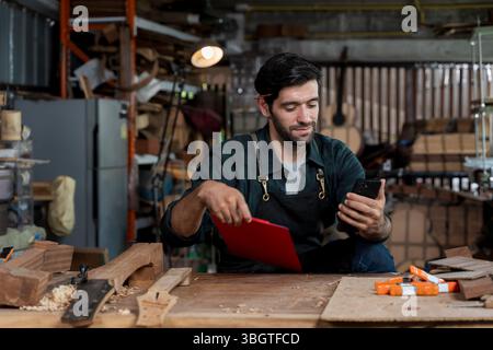 Young male carpenter using smartphone and holding clipboard in woodworking workshop. Concept of modern craftsmanship, multitasking, small business own Stock Photo